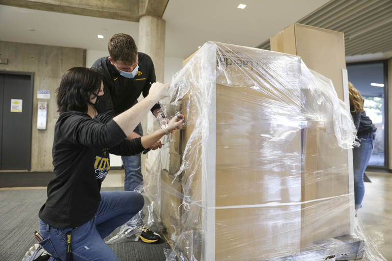 A man and a woman bend over a pallet of boxes and pull off the shrink wrap. 