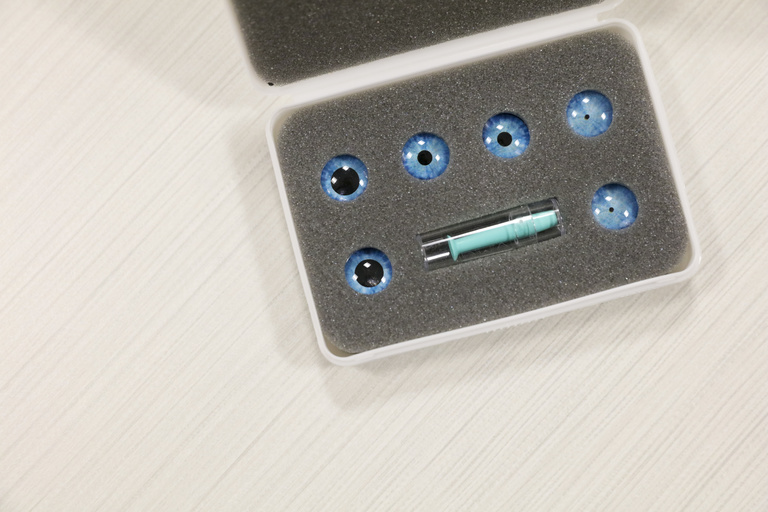 A white plastic case is open on a table. Inside the case is grey foam and six blue circles that look like contact lenses.
